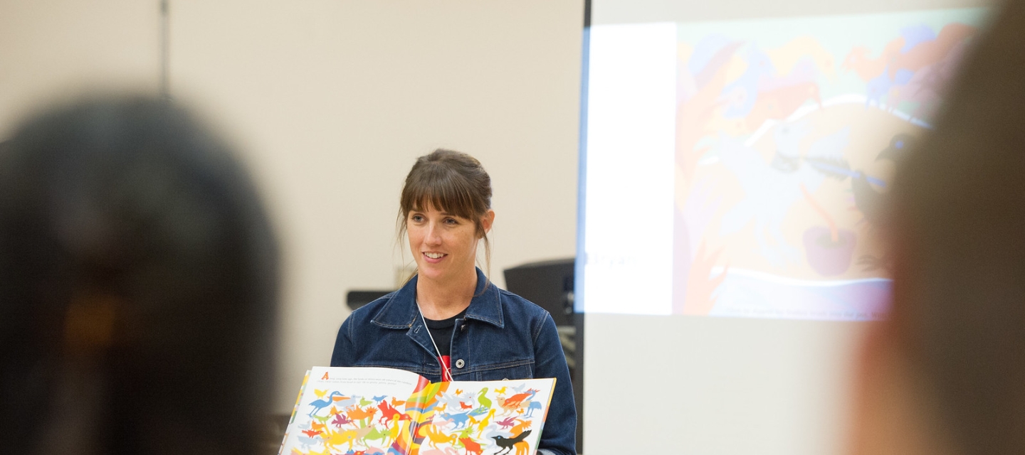 Teacher with children's book stands in front of classroom.